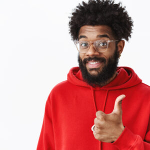 Man showing great gesture being satisfied with awesome quality raising thumb up and smiling friendly in like and approval sign standing delighted over gray background in glasses and red hoodie.